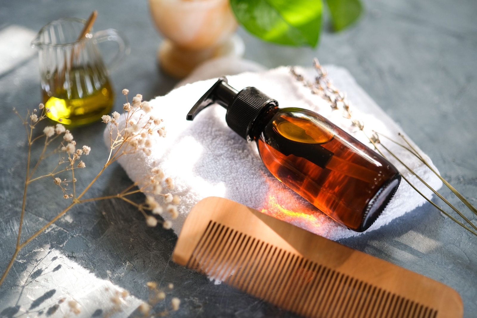 Amber hair oil bottle with a pump dispenser displayed alongside a wooden comb, towel, and natural botanical elements.